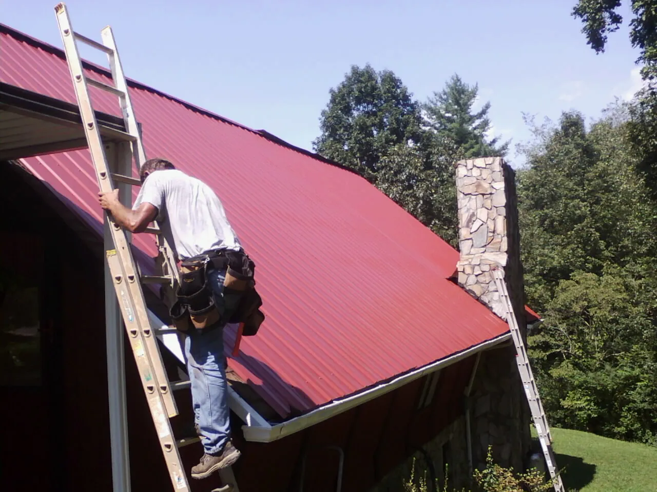 Roofer on a ladder beside a red metal roof and stone chimney — Soffit & Fascia Repair by Roofing Brothers in Verdi